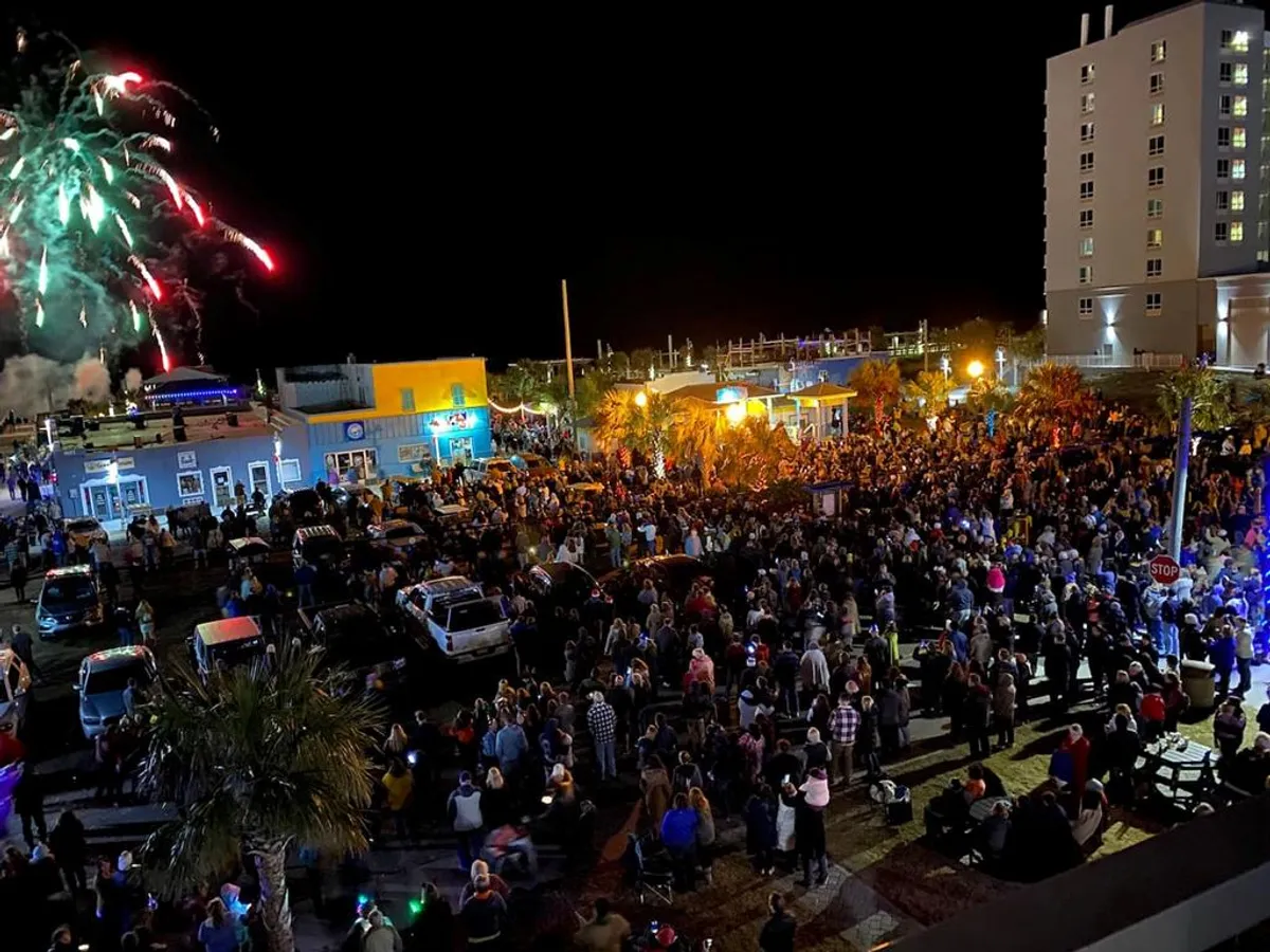 A vibrant exterior scene with a crowd gathered under palm trees, colorful lights illuminating the night, and fireworks bursting in the sky above local buildings. The atmosphere is festive, with people enjoying the lively community spirit and celebration.