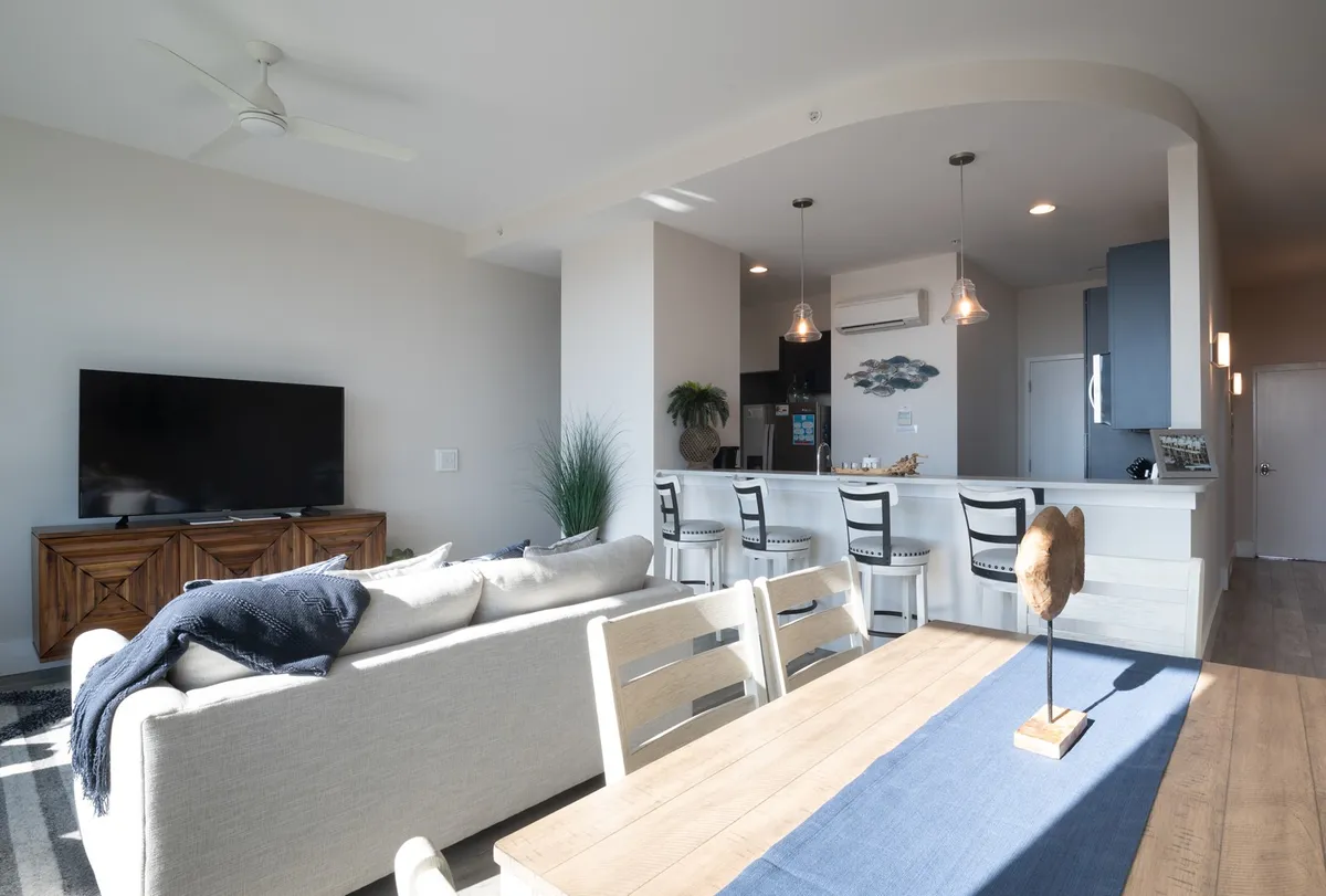 The scene reveals a modern, comfortable living space with a plush sofa, a dining table nearby, a kitchen island, and a wall-mounted television. Tasteful decorative elements, including art on the walls, enhance the welcoming feel. The layout allows for easy flow between lounging, dining, and entertaining.