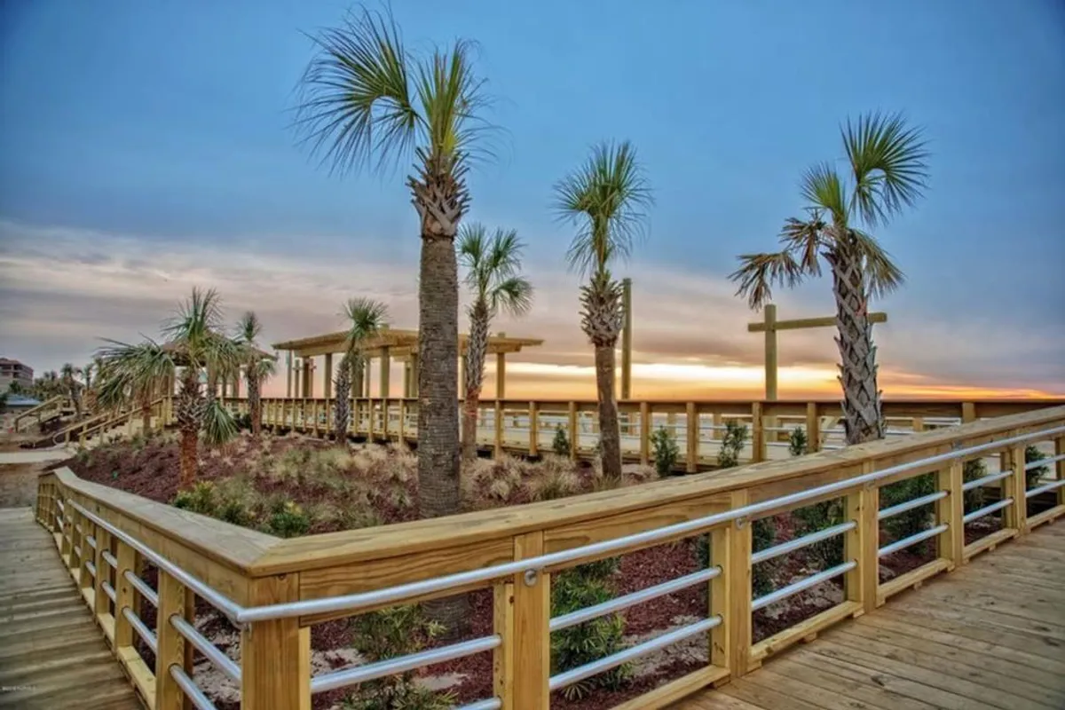 An inviting sunset scene along a wooden walkway lined with palm trees, leading to a viewing platform overlooking the ocean. The sky is painted with warm sunset hues, creating a tranquil and picturesque setting perfect for evening strolls.