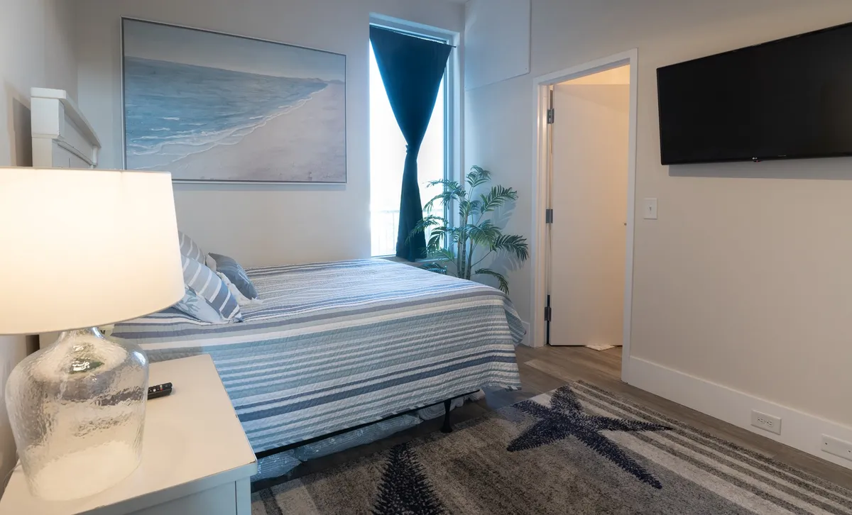 This image features a serene bedroom with a plush bed dressed in striped bedding, a large painting of a beach scene above the headboard, a modern lamp on the bedside table, and a potted plant near the window. The soft lighting, calming decor, and cozy linens create a tranquil and inviting atmosphere.