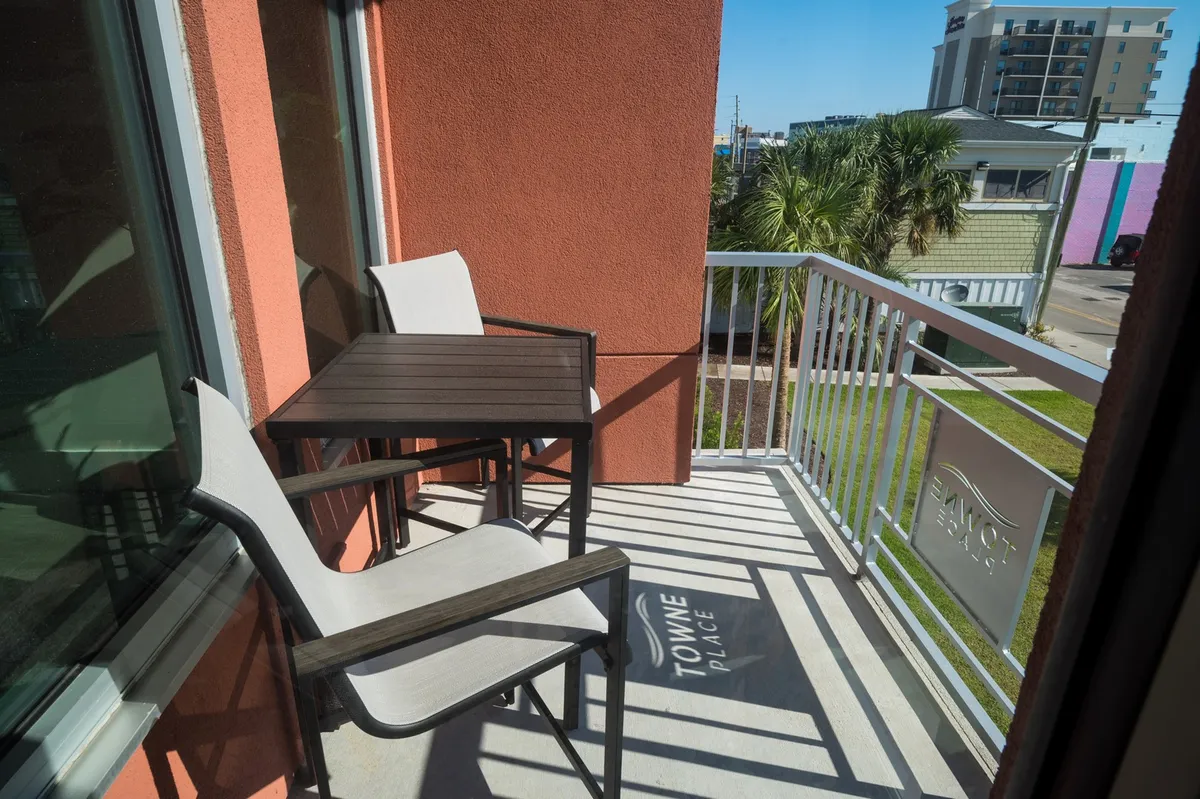 A private balcony set with a small table and chairs overlooks lush palm trees and offers a serene view—perfect for sipping coffee or enjoying the evening ambiance. Ample sunlight and fresh air reinforce the connection to the outdoors and coastal environment.