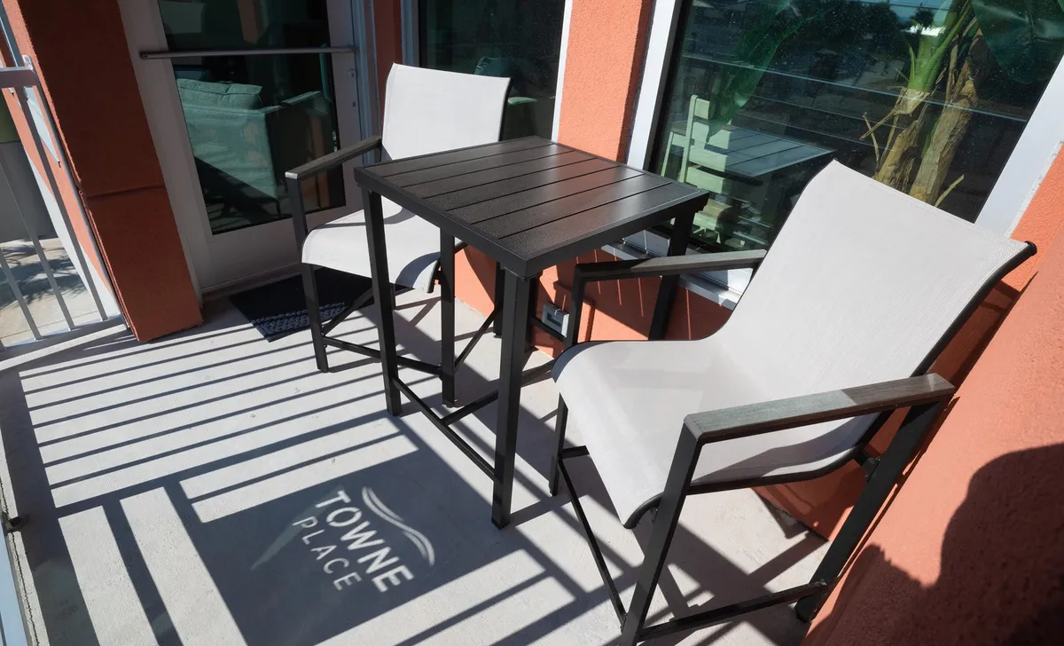 Showing an outdoor table and two chairs with the shadow of a logo cast on the ground, this image evokes the leisurely moments spent enjoying coffee, conversation, or sunset views on the balcony or patio. The bright lighting and casual setup highlight comfort and connection to the outdoors.