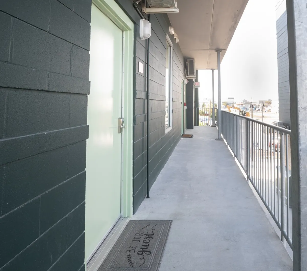 The exterior image centers on a green door with a welcome mat, railings, and hints of the surrounding area. It conveys a sense of both hospitality and security while hinting at the proximity to local attractions. This entrance shot emphasizes the ground-level, gated entry experience.
