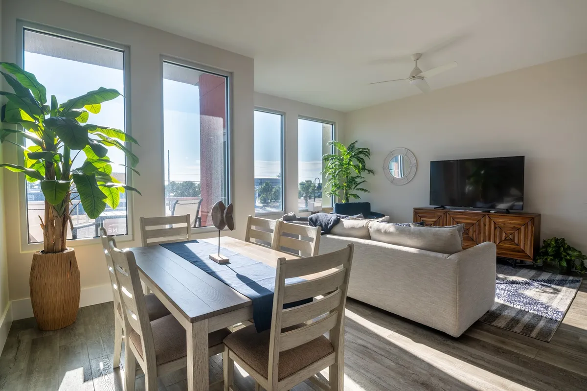 A spacious and bright open-plan living area is depicted, featuring a cozy sofa facing a large television, a stylish dining table set for meals, and expansive windows that flood the space with natural light. Lush potted plants and warm decor give the room a welcoming and relaxed ambiance, ideal for both movie nights and lively gatherings.