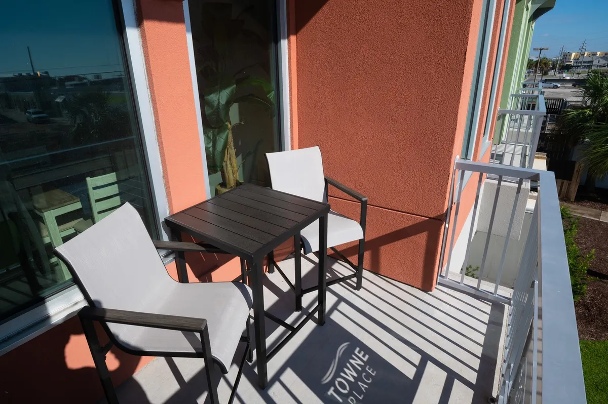 This image features a small outdoor dining table with two chairs set against a colorful wall, ideal for enjoying morning coffee or engaging in relaxed conversation while soaking in the neighborhood atmosphere. The view suggests a comfortable spot to unwind and watch life on the boardwalk.