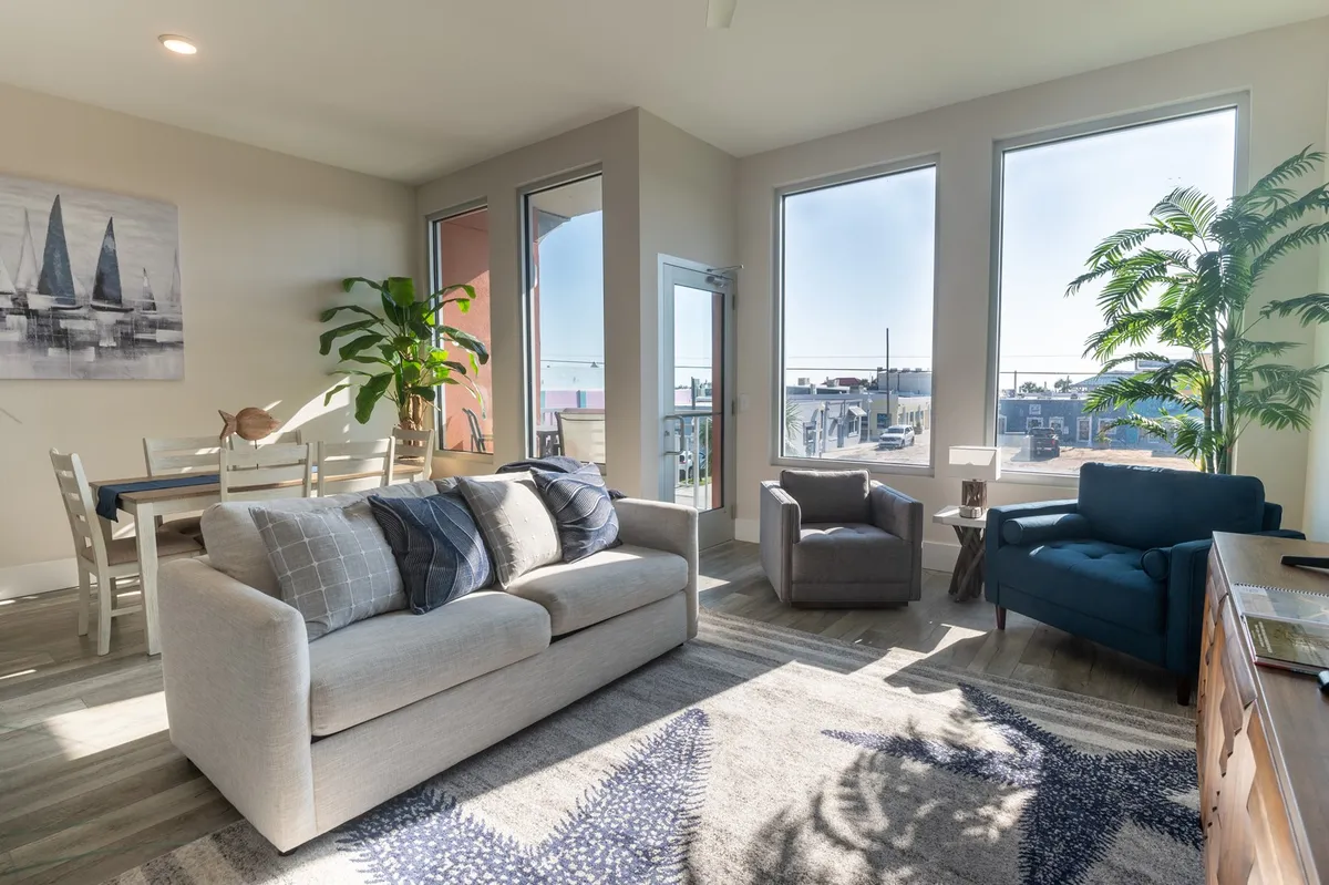 The image showcases a bright, airy living space with expansive windows that bathe the room in natural light. Comfortable seating is arranged for relaxation, with a dining table nearby perfect for meals or casual gatherings. Lush indoor plants add a fresh, inviting touch, and the cheerful decor creates a welcoming atmosphere ideal for unwinding or entertaining.