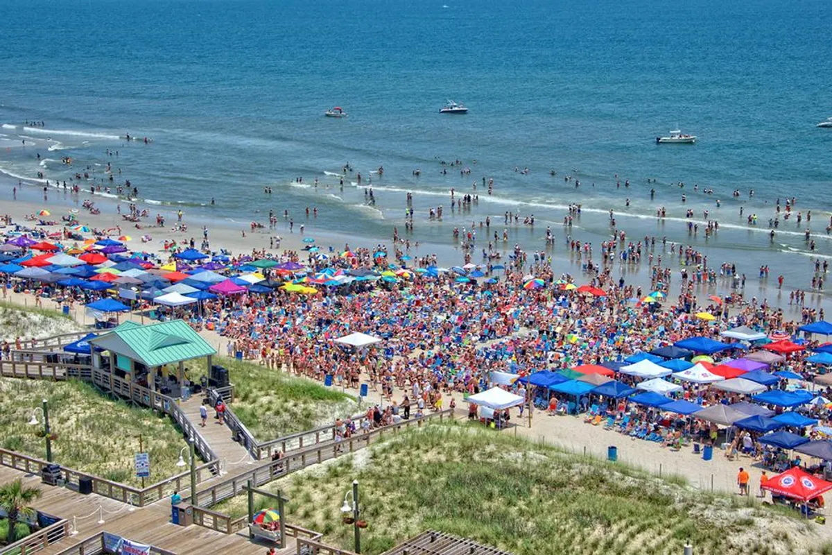 This vibrant exterior shot captures a lively beach scene filled with colorful umbrellas, families, and the ocean waves rolling in. The energy is festive, reflecting sun-filled days of play, socializing, and relaxation right by the water.
