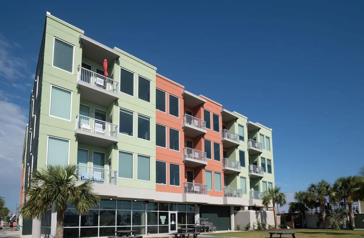 An exterior view of a cheerful, colorful building with balconies and a landscaped area featuring palm trees. The space feels vibrant and welcoming, hinting at the lively atmosphere and social opportunities nearby.