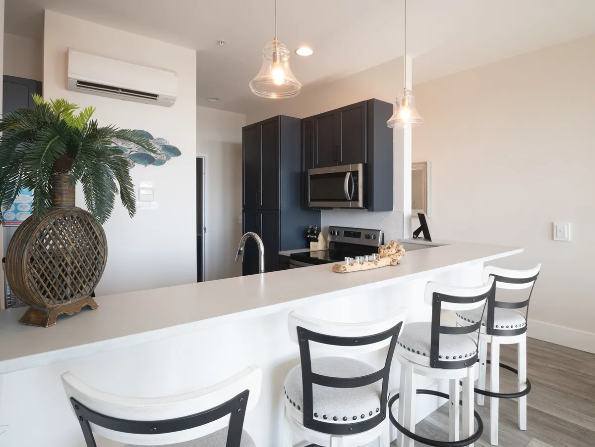 A bright kitchen with a central island, bar stools for countertop seating, a potted plant for a touch of green, and visible appliances. The layout is open, encouraging social interaction while cooking or dining.