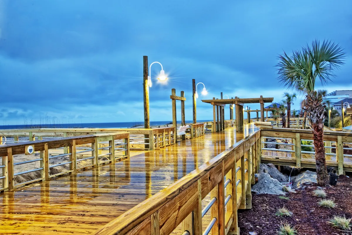 A scenic wooden boardwalk stretches toward the ocean, framed by palm trees and lampposts. The mood is calm and inviting, evoking leisurely strolls and moments of connection by the water.